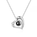 Silver heart-shaped projection necklace with black projection bead, adorned with small clear stones along the heart outline. A photo of a smiling couple is projected below the pendant, while a smaller image of the couple is displayed in the top-right corner. The necklace hangs on a delicate silver chain against a white background.