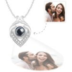 A silver heart-shaped pendant necklace with a central black projection element surrounded by small crystals, displaying a customizable engraved photo below the pendant of a couple smiling and the man kissing the woman’s cheek.