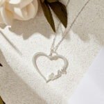 Personalized silver heart-shaped necklace with the name 'Cathy' engraved along the bottom right side, displayed on a textured beige surface with a white flower partially visible in the top left corner, highlighting delicate chain links and elegant script font.