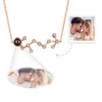 A rose gold constellation-themed pendant necklace with a central black projection element and small crystal accents forming the star pattern, displaying a customizable engraved photo below the pendant of a couple touching foreheads and smiling while holding coffee cups.