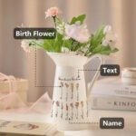 White ceramic pitcher filled with pastel birth flowers, labeled 'Grandma's Garden', featuring names of family members under each flower, arranged neatly on a light-colored table with books and a pink ribbon in the background.
