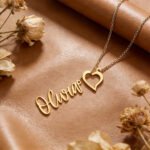 Gold heart name lariat necklace on tan background with dried flowers, featuring a personalised nameplate and delicate heart-shaped design