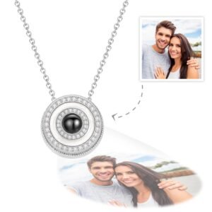 A silver circular pendant necklace with concentric rings of small crystals and a central black projection element, displaying a customizable engraved photo below the pendant of a smiling couple outdoors.
