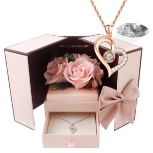 A rose gold heart-shaped pendant necklace with a central black projection element and small crystals along one side, displayed inside a pink gift box with a drawer, surrounded by artificial pink roses, showing a customizable engraved photo of a smiling couple in the top corner.