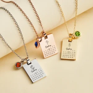 Three rectangular calendar pendant necklaces in silver, rose gold, and gold, each engraved with the name "Sophia," a small flower illustration, and a mini calendar with a heart marking a date, accompanied by a small colored gemstone charm, displayed on delicate chains.