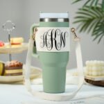 Mint green insulated travel tumbler with handle and cream crossbody strap featuring gold clasps, centered on a white tabletop. The tumbler has a white wrap label displaying ornate black monogram initials “HKN” in decorative script, styled in a clean, modern product photography setup with soft blurred background decor.
