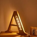 Warm amber-lit decorative LED name lamp shaped like a large letter “A,” with the name “ANDREA” illuminated vertically inside the structure. The lamp sits on a tabletop in a cozy indoor setting, casting a soft golden glow on the wall.
