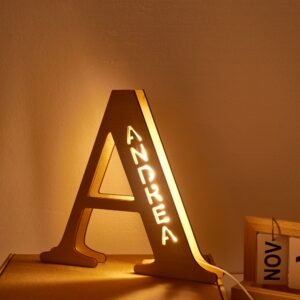 Warm amber-lit decorative LED name lamp shaped like a large letter “A,” with the name “ANDREA” illuminated vertically inside the structure. The lamp sits on a tabletop in a cozy indoor setting, casting a soft golden glow on the wall.