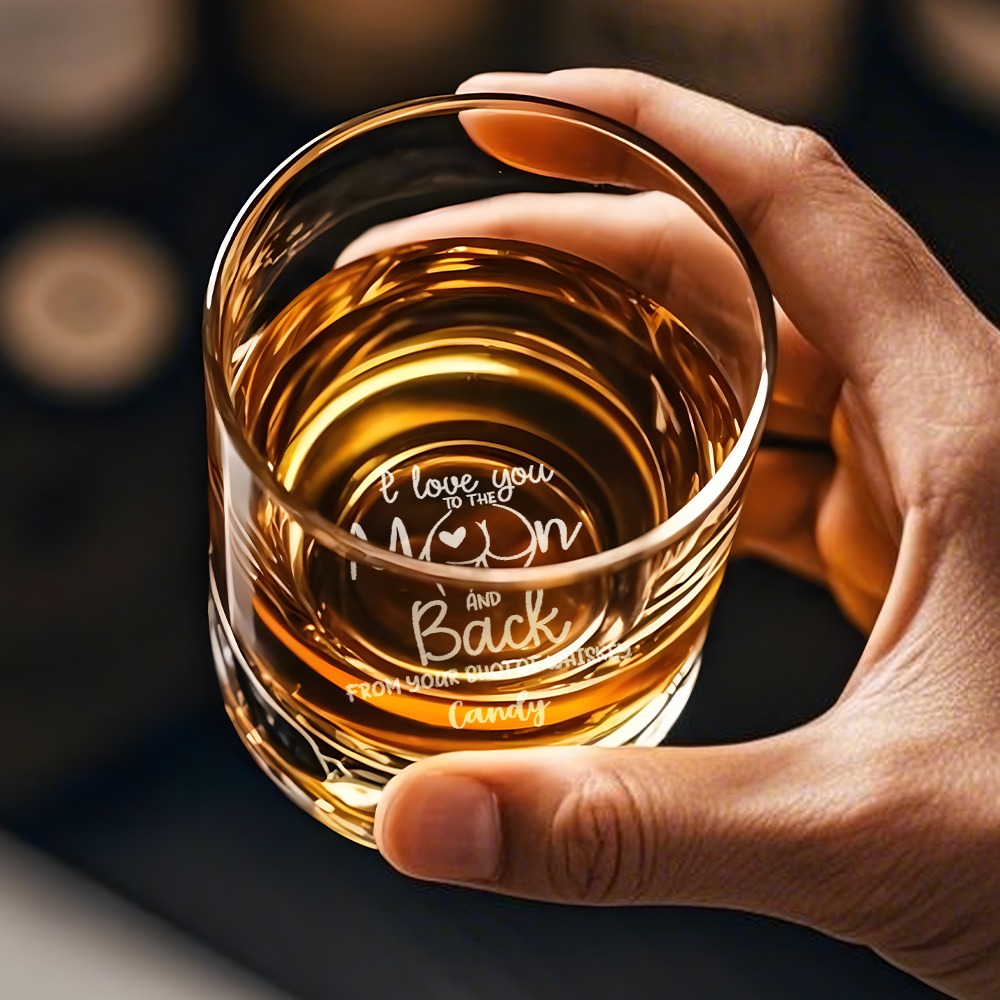 1bb839ab-8a5e-4070-9684-8f027ffed951-0jpeg - Printshop4me.au Close-up of a hand holding a glass of whiskey from above, showing engraved romantic text inside the glass that reads “I love you to the moon and back… From your favourite whiskey Candy,” highlighting a personalized gift message etched into the drinkware.