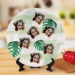 Round ceramic plate displayed on a wooden stand, featuring a repeating collage pattern of a woman’s face with floral crown over a green tropical leaf design background, shown in a warm indoor setting with decorative tomatoes and kitchen props.
