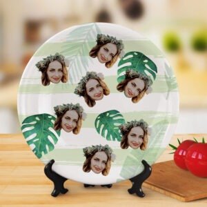 Round ceramic plate displayed on a wooden stand, featuring a repeating collage pattern of a woman’s face with floral crown over a green tropical leaf design background, shown in a warm indoor setting with decorative tomatoes and kitchen props.