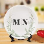 Elegant personalized white ceramic plate featuring the initials “M | N” in the center, surrounded by a delicate green watercolor floral wreath and subtle gold geometric accents near the rim. The plate is displayed on a stand on a wooden surface with a softly blurred kitchen-style background.