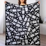 A woman holding up a large custom black-and-white blanket featuring a bold abstract pattern of repeated geometric shapes, stars, and crayon-like icons. The blanket is spread wide to display the full design in a modern indoor room with a clean minimalist background.