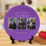 Round personalized commemorative plate displayed on a wooden stand, featuring a purple background design with the text “Celebrating 90 Years Charlie Atkinson” and “October 20 2021,” along with a three-photo timeline layout showing portraits from childhood to adulthood, placed in a warm kitchen-style setting.