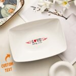 4d61d61a-4bb6-43a3-99cc-5c176e767b0a-0jpeg - Printshop4me.au White rectangular personalized ceramic plate featuring the word “LOVE” with a heart in place of the “O” and red wings extending from both sides. Below it is custom text reading “Michael – 05.10.2022.” An orange “CUSTOM TEXT” label with an arrow points to the personalization area. The plate is styled on a neutral tabletop with decorative items in the background.