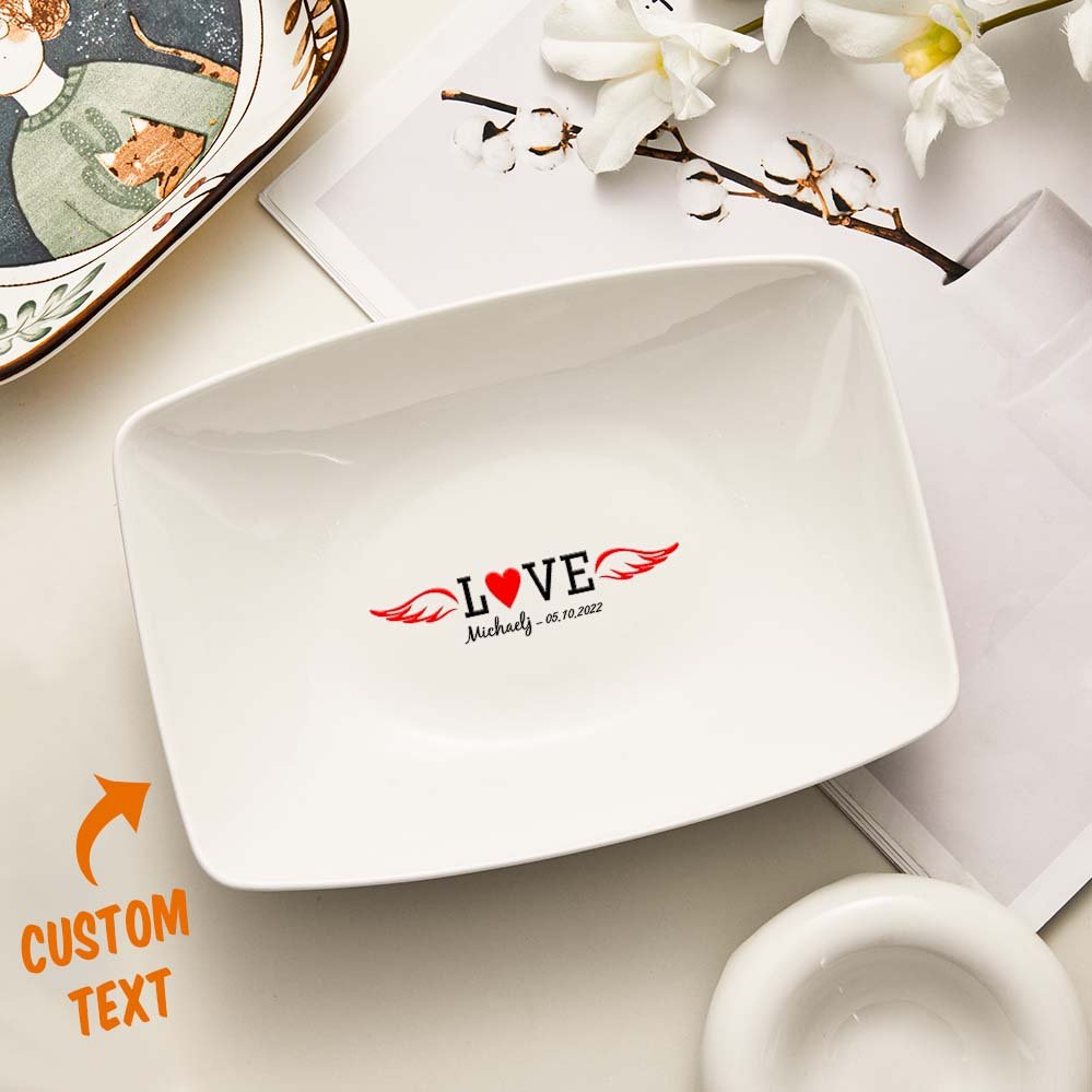 4d61d61a-4bb6-43a3-99cc-5c176e767b0a-0jpeg - Printshop4me.au White rectangular personalized ceramic plate featuring the word “LOVE” with a heart in place of the “O” and red wings extending from both sides. Below it is custom text reading “Michael – 05.10.2022.” An orange “CUSTOM TEXT” label with an arrow points to the personalization area. The plate is styled on a neutral tabletop with decorative items in the background.
