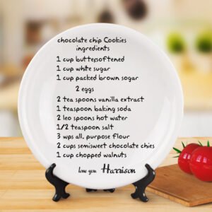 White ceramic decorative plate displayed on a stand featuring handwritten-style recipe text for “chocolate chip Cookies ingredients.” The list includes butter, white sugar, brown sugar, eggs, vanilla extract, baking soda, hot water, salt, all-purpose flour, semisweet chocolate chips, and chopped walnuts. At the bottom, it is signed “love you Harrison.” The plate is placed on a wooden surface with a blurred kitchen-style background and tomatoes on the side.