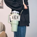 Mint green insulated travel tumbler with handle and cream crossbody strap featuring gold clasps, centered on a white tabletop. The tumbler has a white wrap label displaying ornate black monogram initials “HKN” in decorative script, styled in a clean, modern product photography setup with soft blurred background decor.