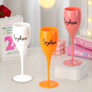 Three personalized champagne flute-style glasses in white, orange, and pink, each printed with the name “Sophia,” displayed on a styled tabletop with soft decorative props in the background, showcasing customizable drinkware options.