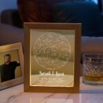 Personalized wooden-framed illuminated wedding keepsake featuring a star map design inside a glowing acrylic panel. The artwork shows “Sarah & Luca” with the message “The Day We Said ‘I Do!’” along with a date and geographic coordinates at the bottom. The frame is placed on a table beside a whiskey glass and a small photo frame, creating a warm romantic home décor setting.