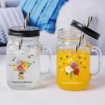 Two personalized mason jar style glass mugs with lids and metal straws, one filled with a white drink and the other with an orange drink. Both feature floral bouquet designs with butterflies and the phrase “Blessed To Be Called Grandma,” set on a bright, clean tabletop background.