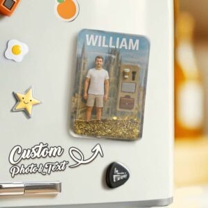 Custom personalized fridge magnet featuring a 3D figurine-style man labeled “WILLIAM,” standing inside a transparent capsule with decorative elements including a coffee cup, flashlight, and bag. The base contains glittery gold particles, giving a floating effect. The design is displayed on a refrigerator surface with playful magnets including a star and food icons, alongside text reading “Custom Photo & Text” with an arrow pointing to the product.