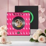 Personalized custom photo music plaque featuring a couple’s photo embedded in a vinyl record-style design on a bright pink background with white lightning bolt pattern. The design shows the song “Beautiful” by Giulio Cercato with a Spotify-style player interface, playback controls, and progress bar. A second black display panel with a green circular record element is placed behind it. The scene is decorated with soft white roses and hanging heart ornaments in a romantic studio setup.