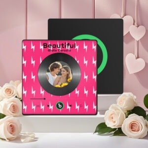 Personalized custom photo music plaque featuring a couple’s photo embedded in a vinyl record-style design on a bright pink background with white lightning bolt pattern. The design shows the song “Beautiful” by Giulio Cercato with a Spotify-style player interface, playback controls, and progress bar. A second black display panel with a green circular record element is placed behind it. The scene is decorated with soft white roses and hanging heart ornaments in a romantic studio setup.
