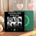 Personalized custom photo music plaque titled “FOREVER LOVE – EDISON & BELLA” featuring a collage of four romantic couple photos in black-and-white. The design is displayed in a Spotify-style music player interface with playback controls, progress bar, and a green circular record element. The plaque has a black background with a green accent border and is set in a warm indoor setting with soft heart decorations and flowers in the background.