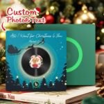 Personalized custom photo and text music plaque featuring a vinyl record-style design with a circular couple photo in the center. The artwork displays the song "All I Want for Christmas Is You" by Mariah Carey, set on a festive winter-themed background with Santa’s sleigh, Christmas trees, stars, and snow effects. A Spotify-style music code and playback controls are shown at the bottom, with a second green display panel in the background.
