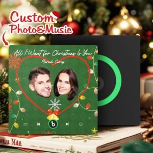 Personalized Christmas-themed custom photo and music plaque featuring a couple’s faces inside a red heart frame on a green festive background. The design shows the song “All I Want for Christmas Is You” by Mariah Carey, with holiday decorations including lights, bells, and a snowflake ornament. A Spotify-style music interface and playback controls appear at the bottom, with a secondary display panel in the background on a Christmas-themed setting.