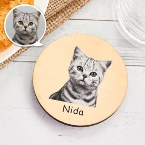 Round wooden personalized pet plaque featuring a grayscale portrait of a kitten printed in the center, with the name “Nida” written below. A small speech-bubble style inset shows a close-up of the cat’s face in the top-left corner. The plaque is placed on a light wooden surface with decorative table elements around it.