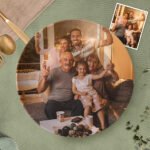 Personalized circular photo plaque featuring a warm family portrait of three generations sitting together in a cozy living room. The image shows smiling grandparents, parents, and a child posing happily, with soft golden lighting enhancing the scene. A small inset photo appears in the top-right corner showing another family group shot. The plaque is displayed on a textured green surface with decorative table elements, creating a sentimental custom family keepsake.