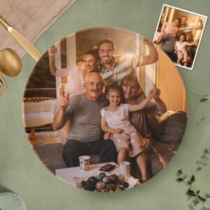 Personalized circular photo plaque featuring a warm family portrait of three generations sitting together in a cozy living room. The image shows smiling grandparents, parents, and a child posing happily, with soft golden lighting enhancing the scene. A small inset photo appears in the top-right corner showing another family group shot. The plaque is displayed on a textured green surface with decorative table elements, creating a sentimental custom family keepsake.