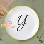 Top-down view of a white ceramic plate with a yellow rim placed on a green table setting. A large stylized letter “Y” is printed in the center of the plate, with orange text reading “your Text here” and an arrow pointing toward the customizable area, indicating space for personalization. The scene includes partial cutlery and fabric napkin for styling.
