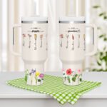 Two personalized insulated tumblers displayed side by side on a countertop, labeled “First mom” and “now grandma,” decorated with delicate floral illustrations and handwritten-style names of flowers. Each tumbler has a lid and straw, representing a sentimental gift design celebrating motherhood and grandmotherhood.