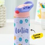 Personalized kids' stainless steel water bottle with a lavender and pink flip-top straw lid and carry handle, featuring a colorful space-themed design with planets, stars, and the sun. The bottle is customized with the name "Lulia" and displayed on a desk with a "Custom Name" label.