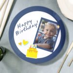 Personalized birthday ceramic plate with a navy blue rim featuring the text “Happy Birthday!” in a handwritten-style font. The design includes small heart and cake illustrations and a tilted photo frame of a young child eating with a spoon. The plate is placed on a table setting with cutlery and a soft fabric background, creating a festive custom gift presentation.