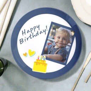 Personalized birthday ceramic plate with a navy blue rim featuring the text “Happy Birthday!” in a handwritten-style font. The design includes small heart and cake illustrations and a tilted photo frame of a young child eating with a spoon. The plate is placed on a table setting with cutlery and a soft fabric background, creating a festive custom gift presentation.