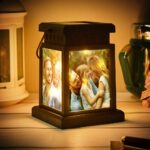 Warm glowing personalized photo lantern placed on a wooden surface, featuring illuminated panels with family photographs on each side. The lantern emits a soft golden light showcasing smiling parents and child in an outdoor setting. The scene is styled with ambient lighting and home décor items in the background, creating a cozy decorative gift atmosphere.