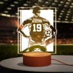 XHD3721-2jpg - Printshop4me.au Glowing personalized LED sports lamp featuring a football player silhouette with the name “BENZEMA” and number “19” on the back of a jersey design. The lamp emits warm golden light from a rectangular acrylic panel mounted on a round base, placed in a stadium-themed background with blurred seating and pitch lighting.