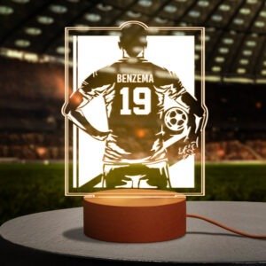 Glowing personalized LED sports lamp featuring a football player silhouette with the name “BENZEMA” and number “19” on the back of a jersey design. The lamp emits warm golden light from a rectangular acrylic panel mounted on a round base, placed in a stadium-themed background with blurred seating and pitch lighting.