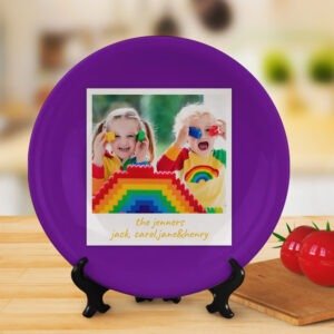 Round personalized photo plate on a wooden stand, featuring a purple background and a central square photo of two children playing with colorful blocks and a rainbow design, with handwritten-style text reading “the jenners jack, carol, jane & henry,” displayed in a warm indoor kitchen setting.