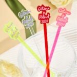 a9c3fa9f-3c64-43fc-ad44-1f84228f8224-0jpeg - Printshop4me.au Colorful personalized drink stirrers placed in a glass bowl, featuring names “Octavia & Patrick 2024” in different styles and colors including red, pink, neon green, and silver. The sticks are arranged like party decorations, suggesting custom wedding or celebration drink accessories.