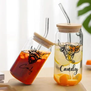 Two personalized glass tumblers with wooden lids and metal straws placed on a wooden surface. One tumbler is tilted and filled with a red iced drink labeled “Candy” with a Highland cow illustration, while the other contains a light yellow drink with the same cow design and matching “Candy” text, showcasing a custom drinkware set.
