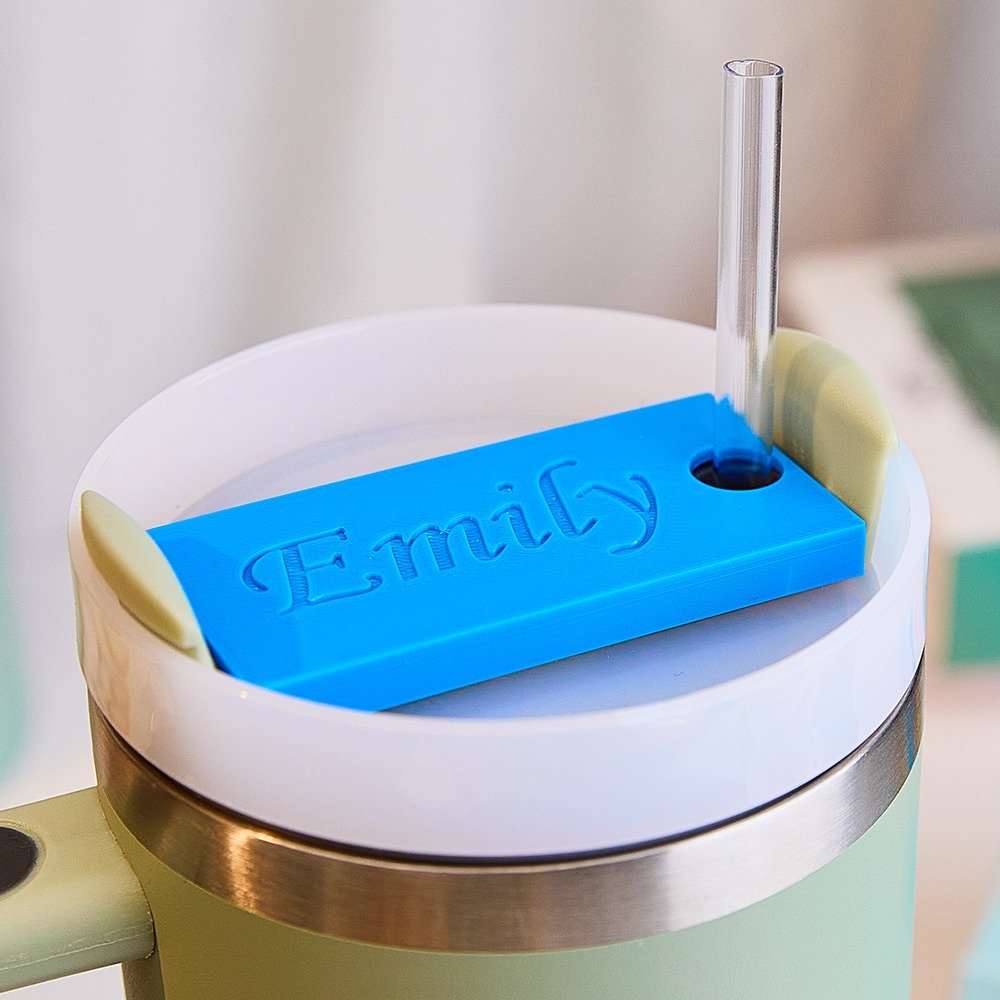 e990e8c8-06b5-467f-b5e3-2064fc96ac7c-0jpeg - Printshop4me.au Close-up of a personalized tumbler lid insert with a bright blue rectangular name plate engraved “Emily,” featuring a straw hole and placed inside a white insulated travel mug lid with a clear straw inserted.