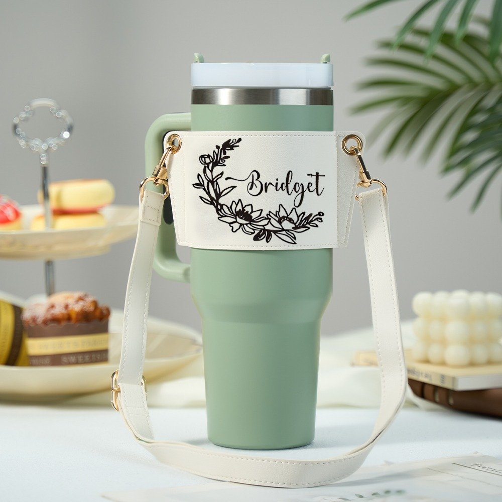 ff84911d-2e81-4dd5-a6e9-6b8e9f5e4de9-0jpeg - Printshop4me.au Mint green insulated travel tumbler with a cream crossbody strap and gold hardware, featuring a white wrap label decorated with a black floral wreath design and the name “Bridget” in script font, styled on a clean tabletop with soft decorative background elements.